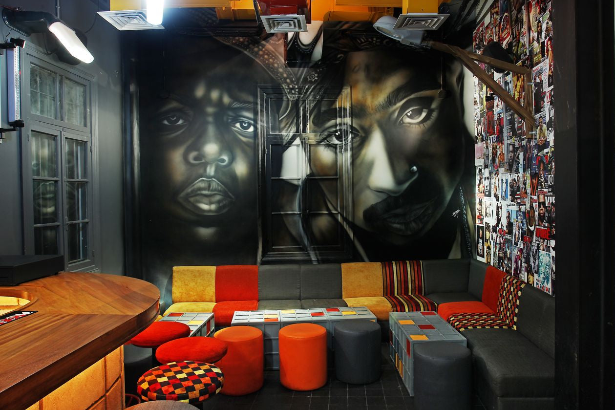 A room with a large canvas artwork featuring a person's face, with a mix of furniture including bean bags and ottomans in an L-shaped arrangement, in a setting that resembles a home interior.